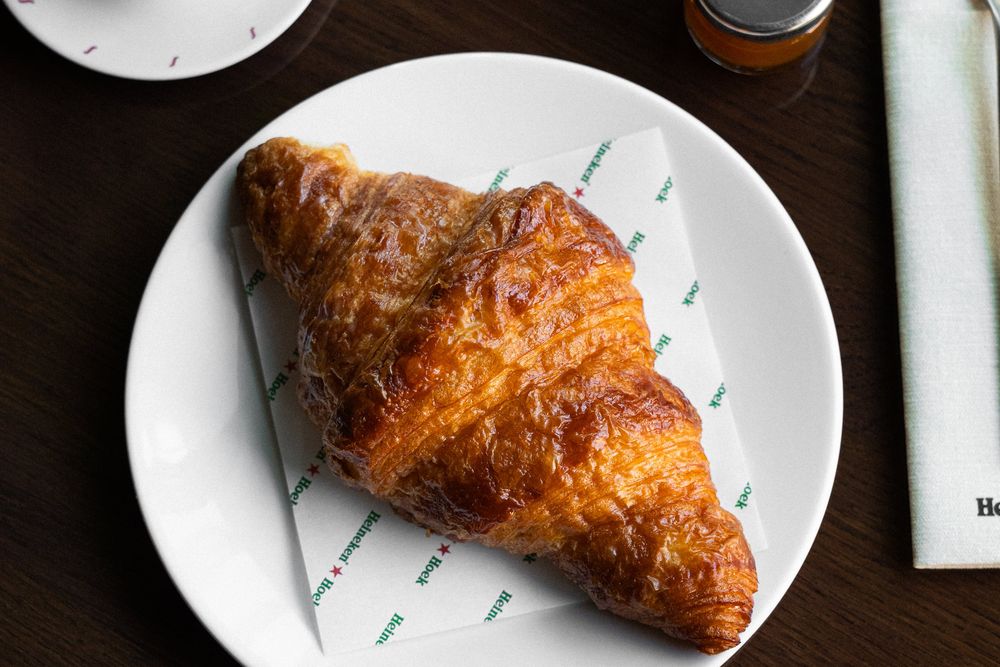 A butter croissant on a Heineken Hoek napkin on a white plate, with a small jar of jam on a dark wooden table